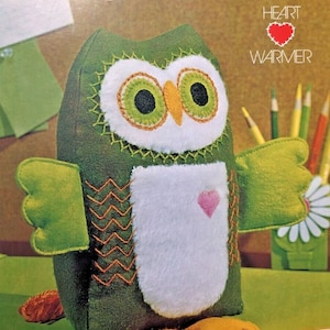 May include: A green felt owl with large eyes, a white belly, and a pink heart on its chest. The owl is labeled "HOOT the owl HEART WARMER".