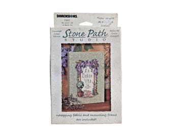 Op de afbeelding: Een Dimensions Stone Path Studio telpatroon kruissteekpakket met een ontwerp genaamd La Dolce Vita The Good Life. De verpakking toont een afgewerkt project met een bloemenboog, een potplant en tekst, inclusief een montageraam.