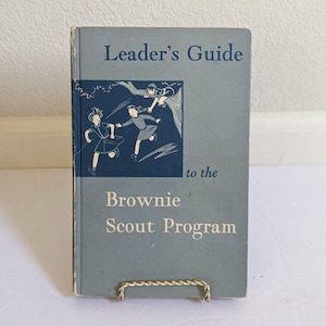 May include: A blue book with the title "Leader's Guide to the Brownie Scout Program". The cover features an illustration of three girls in uniform playing.