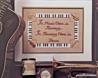 Op de afbeelding: Een ingelijst kruissteekkunstwerk met de tekst "In Music there is Harmony... In Harmony there is Peace." Het kunstwerk is omgeven door pianotoetsen en muziekinstrumenten. Een mok, sleutelhanger, gitaar en trompet zijn ook zichtbaar.