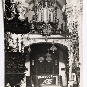 May include: A black and white photo of the interior of a church. The photo shows a large pipe organ, a wooden door with intricate carvings, and rows of pews. The text "TAXCO. GRO." is visible on the door.
