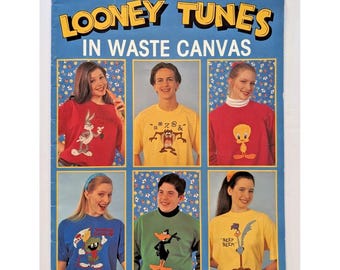 Op de afbeelding: Een Leisure Arts Looney Tunes in Waste Canvas knutselfolder, nummer 2564, met zes foto's van personen die shirts dragen met geborduurde Looney Tunes-personages, waaronder Bugs Bunny, Taz, Tweety, Marvin de Marsmannetje, Daffy Duck en Road Runner.