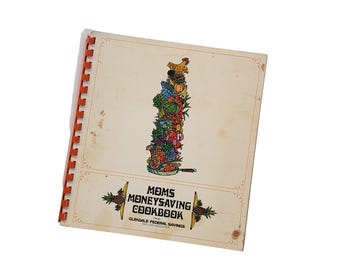 Op de afbeelding: Vintage kookboek getiteld "Moms Moneysaving Cookbook" met een spiraalbinding. De omslag toont een kleurrijke illustratie van opgestapeld fruit en groenten bekroond met een kip. Het kookboek is van Glendale Federal Savings.