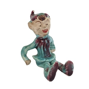 May include: A vintage ceramic figurine of a seated elf with a smiling face. The elf wears a teal outfit with burgundy accents on the chest and feet. The elf's hat and hair are also burgundy. The figurine has a cream-colored face.