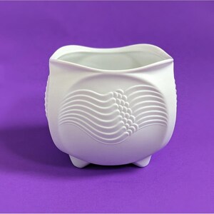 May include: A white ceramic planter with a unique design. The planter has a rounded shape with three small feet and a wavy top edge. The front features an embossed pattern of curved lines and small circles. The background is a solid purple.