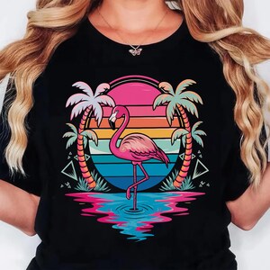 May include: Black t-shirt featuring a pink flamingo standing in water, set against a sunset backdrop with palm trees. The design includes vibrant colors like pink, orange, blue, and green, creating a tropical theme.