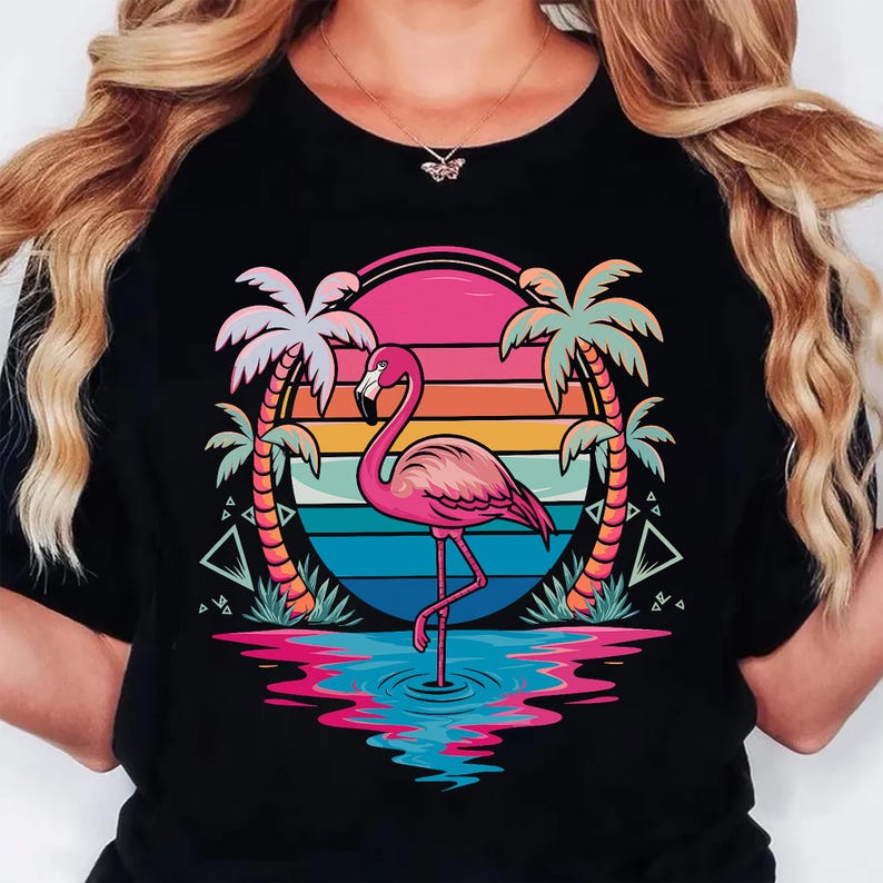May include: Black t-shirt featuring a pink flamingo standing in water, set against a sunset backdrop with palm trees. The design includes vibrant colors like pink, orange, blue, and green, creating a tropical theme.