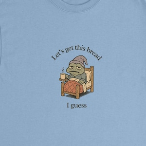 May include: Light blue t-shirt featuring a cartoon frog in a nightcap, sitting in a bed and holding a mug. The text "Let's get this bread" and "I guess" are printed on the shirt.