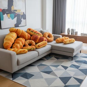 May include: A collection of plush bread-shaped pillows, including baguettes, croissants, and hot dogs, arranged on a gray sofa. The pillows are various shades of brown and yellow, resembling baked goods. A geometric patterned rug is in the foreground.