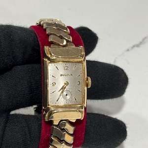May include: A gold-coloured Bulova wristwatch with a rectangular face and a linked band. The watch face is silver with black numerals and hands. The watch is displayed on a red velvet surface.