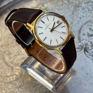 May include: A vintage gold-toned wristwatch with a white face and a brown leather band. The watch features a date display at the 3 o'clock position and is displayed on a clear stand. The watch has the text "Eterna-Matic" on the face.