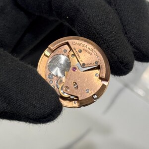 May include: A close-up of a vintage Omega Watch Co. watch movement. The gold-toned mechanism features intricate gears, jewels, and the text "OMEGA WATCH CO SWISS SEVENTEEN 17 JEWELS". The watch movement is held by a black gloved hand.