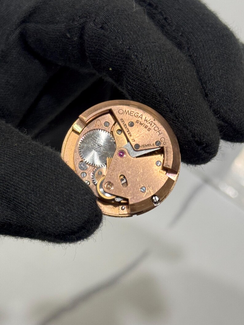 May include: A close-up of a vintage Omega Watch Co. watch movement. The gold-toned mechanism features intricate gears, jewels, and the text "OMEGA WATCH CO SWISS SEVENTEEN 17 JEWELS". The watch movement is held by a black gloved hand.