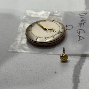 May include: A vintage Omega watch with a gold-tone case and hands, on a plastic bag. The watch face is off-white with black numerals. A separate gold-tone crown is also visible.