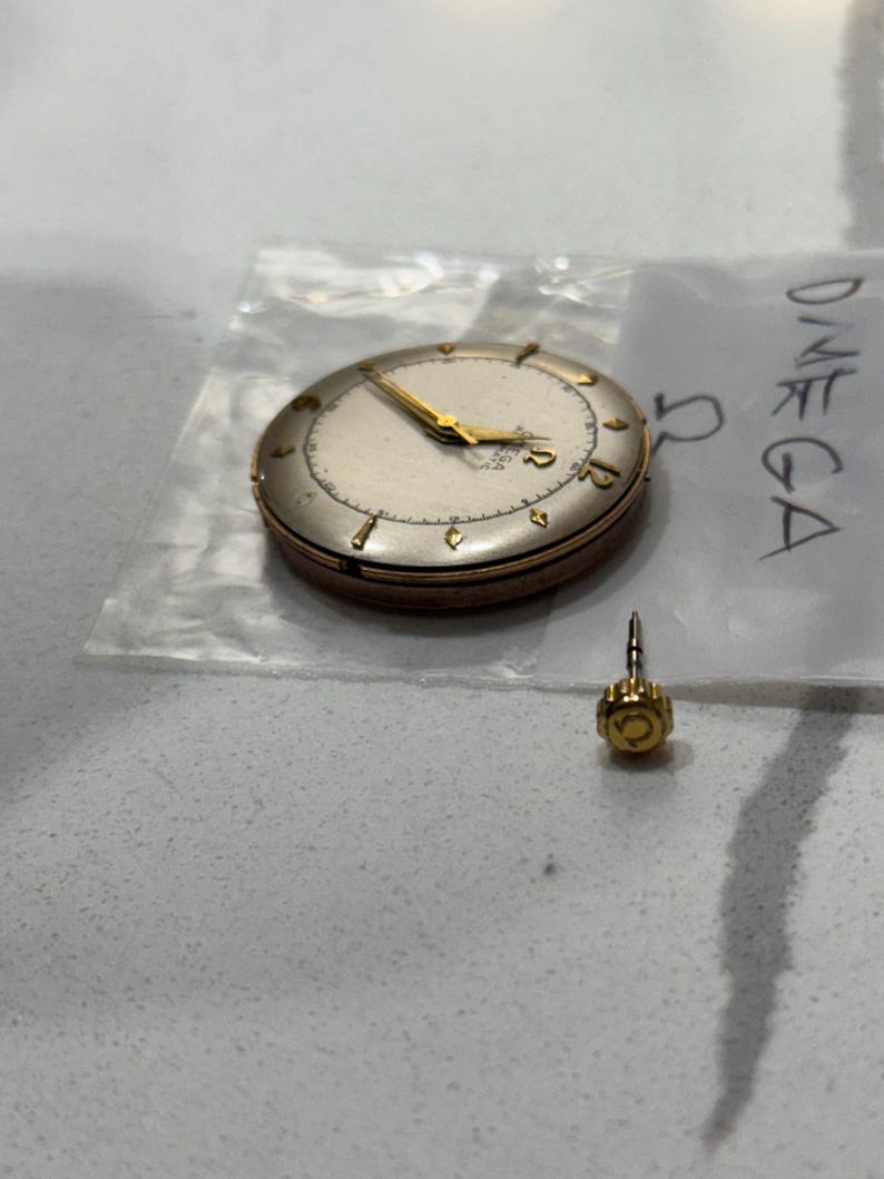 May include: A vintage Omega watch with a gold-tone case and hands, on a plastic bag. The watch face is off-white with black numerals. A separate gold-tone crown is also visible.