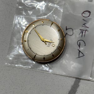 May include: A vintage Omega watch face with a silver dial, gold hands, and gold hour markers. The watch face is round with a gold-coloured bezel. The Omega logo and text are visible on the dial. The watch face is in a clear plastic bag.