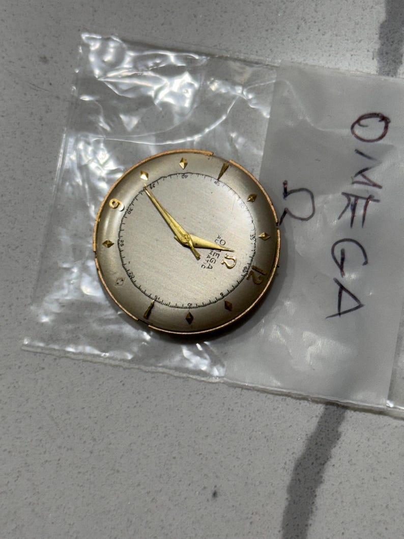 May include: A vintage Omega watch face with a silver dial, gold hands, and gold hour markers. The watch face is round with a gold-coloured bezel. The Omega logo and text are visible on the dial. The watch face is in a clear plastic bag.