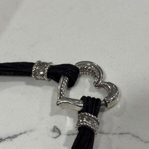 K&ouml;nnte beinhalten: Ein schwarzes Kordelarmband mit einem silbernen herzf&ouml;rmigen Verschluss. Der Verschluss weist aufwendige Details auf, und die Kordel ist mit dekorativen silbernen Akzenten versehen. Das Armband wird vor wei&szlig;em Hintergrund pr&auml;sentiert.