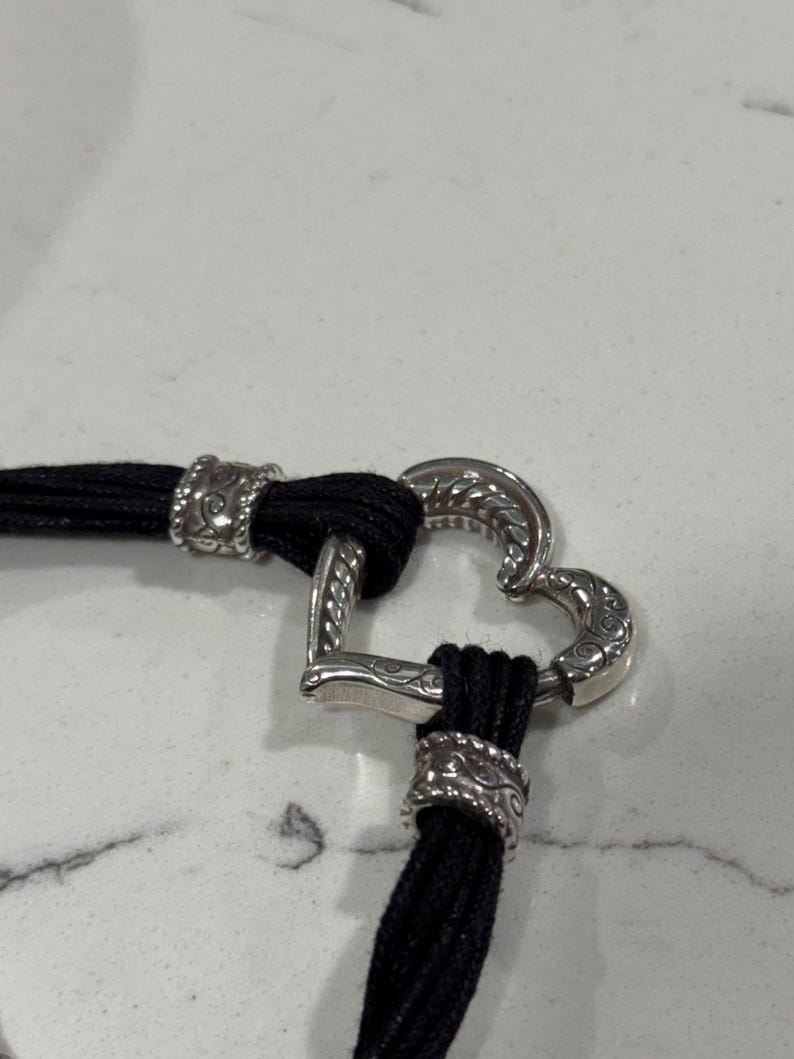K&ouml;nnte beinhalten: Ein schwarzes Kordelarmband mit einem silbernen herzf&ouml;rmigen Verschluss. Der Verschluss weist aufwendige Details auf, und die Kordel ist mit dekorativen silbernen Akzenten versehen. Das Armband wird vor wei&szlig;em Hintergrund pr&auml;sentiert.