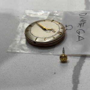 May include: A vintage Omega watch face with gold hands and hour markers. The watch is in a clear plastic bag with the word "OMEGA" written on it. A separate gold watch crown is also visible.