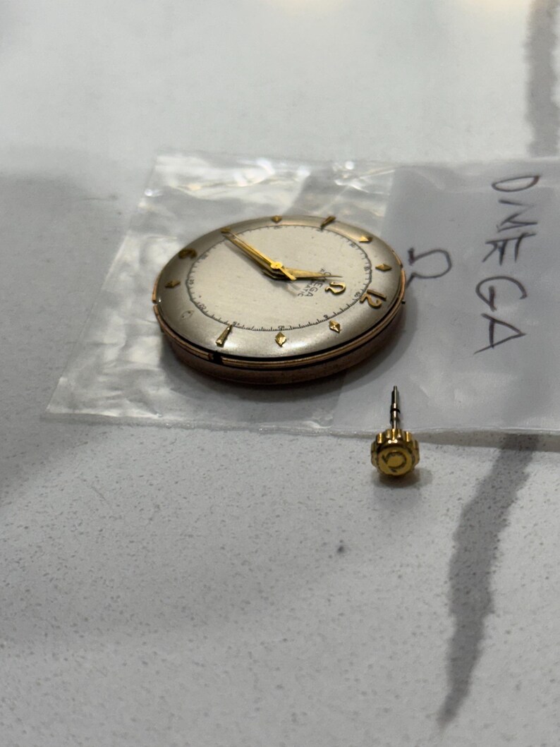 May include: A vintage Omega watch face with gold hands and hour markers. The watch is in a clear plastic bag with the word "OMEGA" written on it. A separate gold watch crown is also visible.