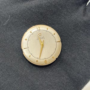 May include: A vintage gold Omega Automatic watch face with a cream dial. The watch has gold hands and hour markers, with the Omega logo and text at the centre. The watch face is set against a dark background.
