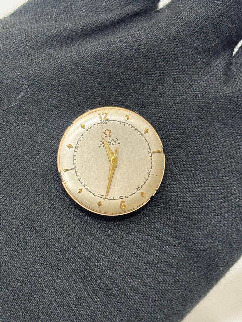 May include: A vintage gold Omega Automatic watch face with a cream dial. The watch has gold hands and hour markers, with the Omega logo and text at the centre. The watch face is set against a dark background.