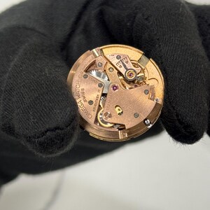 May include: Close-up of a vintage Omega watch movement. The intricate mechanism features gold-toned gears, a balance wheel, and ruby jewels. The watch movement is marked with "Omega Watch Co. Swiss Seventeen 17 Jewels".