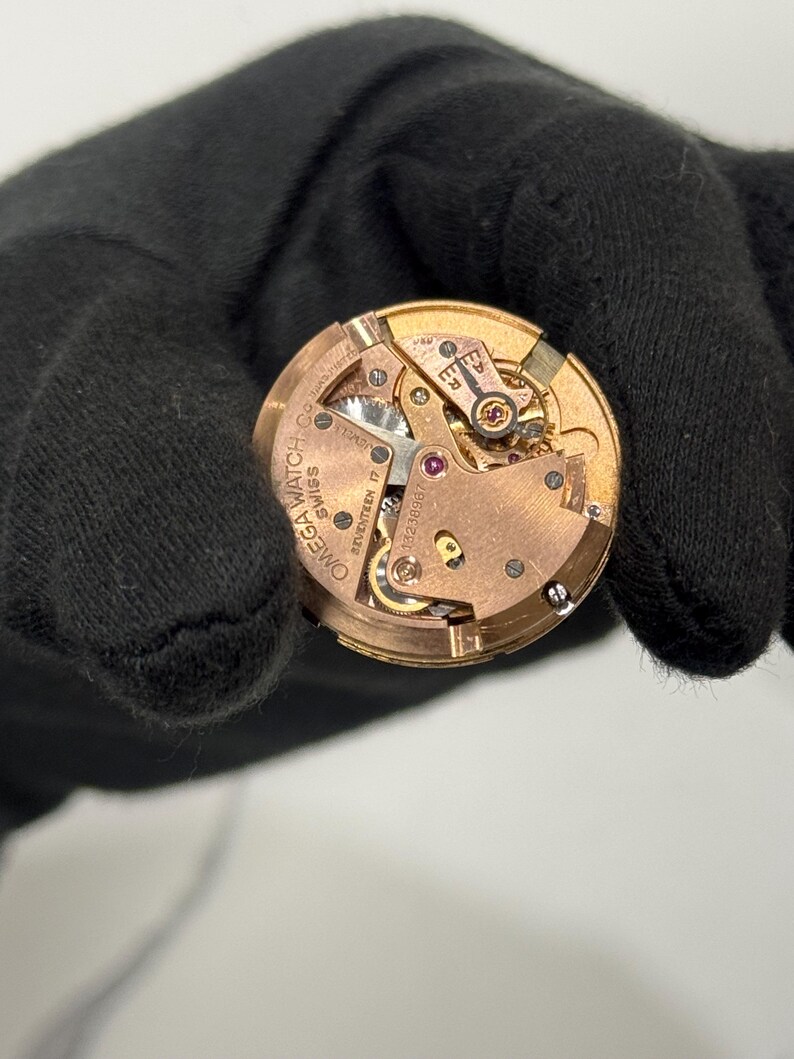 May include: Close-up of a vintage Omega watch movement. The intricate mechanism features gold-toned gears, a balance wheel, and ruby jewels. The watch movement is marked with "Omega Watch Co. Swiss Seventeen 17 Jewels".