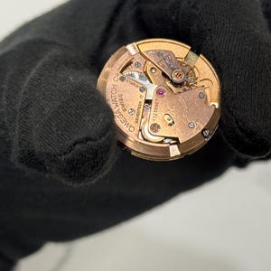 May include: A detailed view of an Omega watch movement, showcasing its internal components. The gold-coloured mechanism includes gears, screws, and a balance wheel. The watch movement is inscribed with "Omega Watch Co. Swiss" and "Seventeen 17 Jewels".