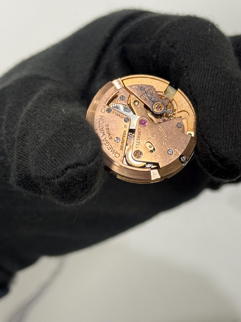 May include: A detailed view of an Omega watch movement, showcasing its internal components. The gold-coloured mechanism includes gears, screws, and a balance wheel. The watch movement is inscribed with "Omega Watch Co. Swiss" and "Seventeen 17 Jewels".