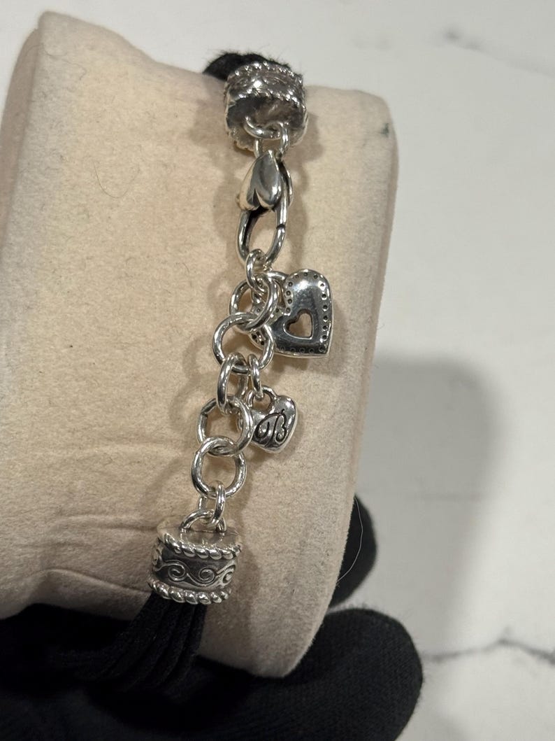 K&ouml;nnte beinhalten: Silberfarbenes Charm-Armband mit herzf&ouml;rmigen Charms und dekorativen Endkappen. Das Armband hat eine schwarze Quaste und wird auf einer neutralfarbenen Oberfl&auml;che pr&auml;sentiert. Die Charms umfassen ein Herz mit einem Ausschnitt und ein kleineres Herz mit Initialen.