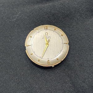 Könnte beinhalten: Eine Vintage Omega Automatic Armbanduhr mit goldfarbenem Gehäuse und silbernem Zifferblatt. Die Uhr hat goldene Zeiger und Stundenmarkierungen, mit dem Omega-Logo und Text sichtbar. Die Uhr ist vor dunklem Hintergrund.