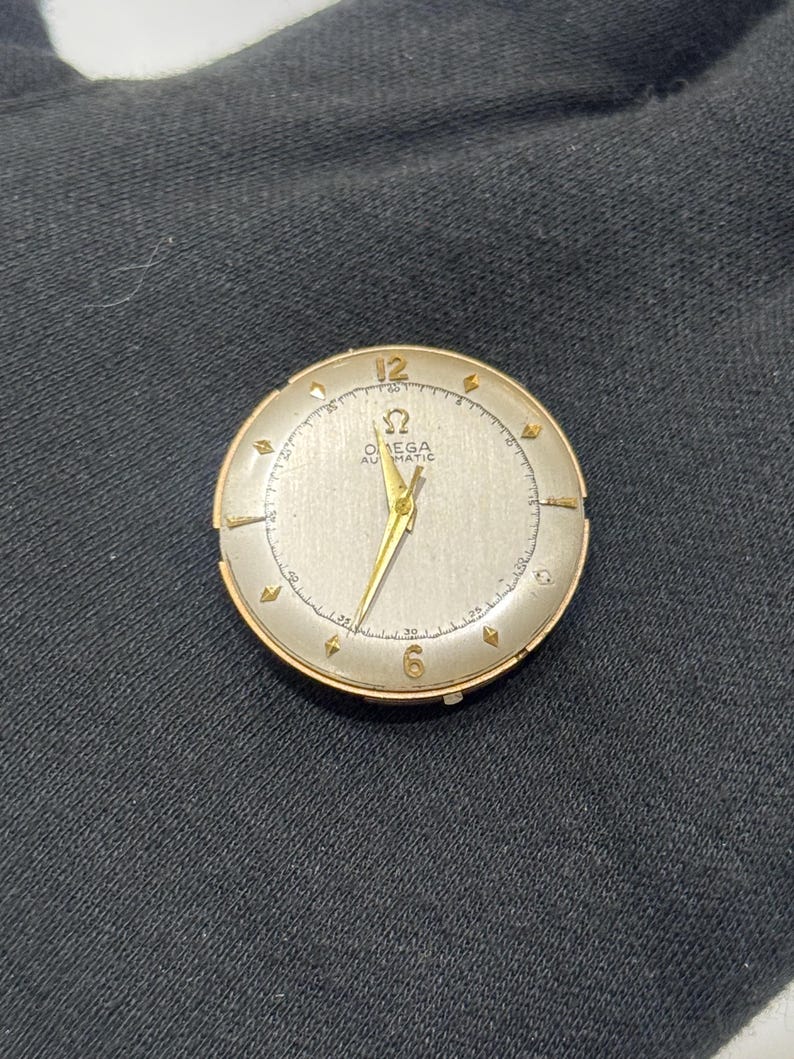 May include: A vintage Omega Automatic wristwatch. It has a gold-tone case and a silver dial. The watch features gold hands and hour markers, with the Omega logo and text visible. The watch is set against a dark background.