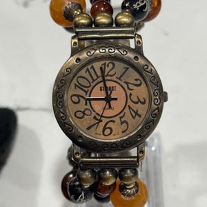 May include: A vintage-style watch with a bronze-colored face and band. The watch face features large, easy-to-read numbers and the brand name "Giande". The band is adorned with amber and brown beads. The watch is a unique accessory.