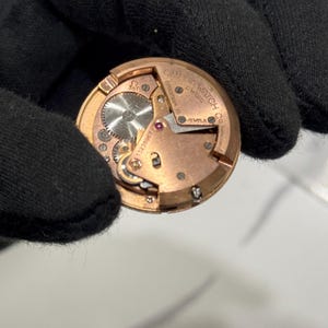 May include: A close-up shot of a vintage Omega watch movement. The gold-coloured mechanism displays intricate gears, a ruby jewel, and the brand name. The watch movement is set against a black background, highlighting the detailed craftsmanship.
