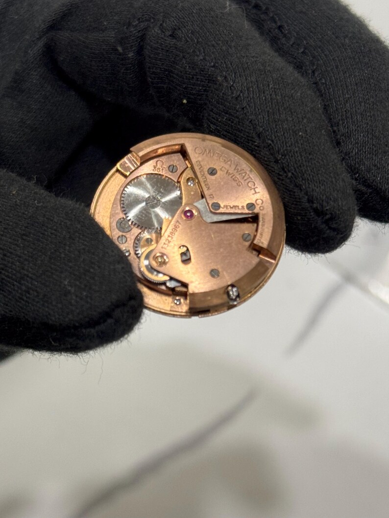 May include: A close-up shot of a vintage Omega watch movement. The gold-coloured mechanism displays intricate gears, a ruby jewel, and the brand name. The watch movement is set against a black background, highlighting the detailed craftsmanship.