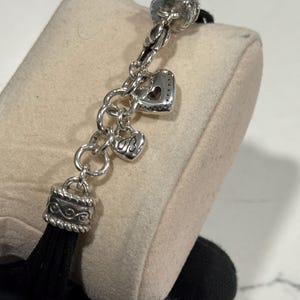 K&ouml;nnte beinhalten: Silberfarbenes Charm-Armband mit herzf&ouml;rmigen Charms und einer schwarzen Quaste. Das Armband verf&uuml;gt &uuml;ber einen dekorativen Verschluss und mehrere Herz-Charms, eines mit einem herzf&ouml;rmigen Ausschnitt. Die Quaste ist schwarz und h&auml;ngt an einer dekorativen silbernen Kappe.