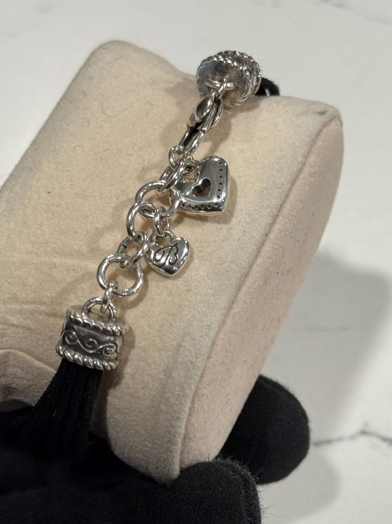 K&ouml;nnte beinhalten: Silberfarbenes Charm-Armband mit herzf&ouml;rmigen Charms und einer schwarzen Quaste. Das Armband verf&uuml;gt &uuml;ber einen dekorativen Verschluss und mehrere Herz-Charms, eines mit einem herzf&ouml;rmigen Ausschnitt. Die Quaste ist schwarz und h&auml;ngt an einer dekorativen silbernen Kappe.
