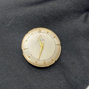 May include: A vintage gold-toned Omega Automatic wristwatch with a silver face. The watch has gold hands and hour markers, with the numbers 12 and 6 visible. The watch is round and appears to be in good condition.