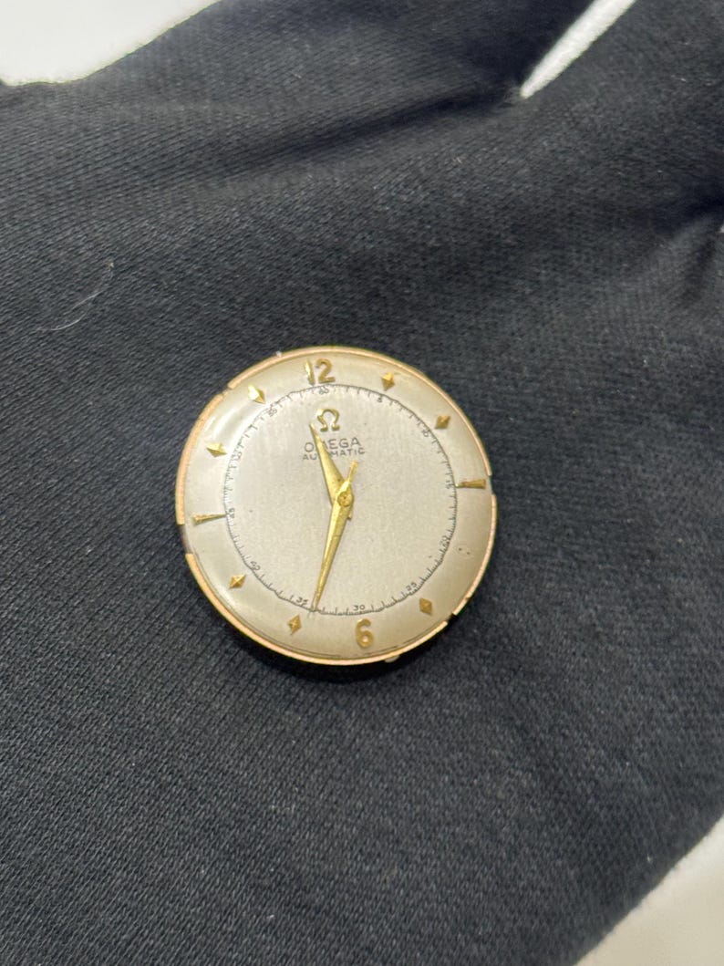 May include: A vintage gold-toned Omega Automatic wristwatch with a silver face. The watch has gold hands and hour markers, with the numbers 12 and 6 visible. The watch is round and appears to be in good condition.