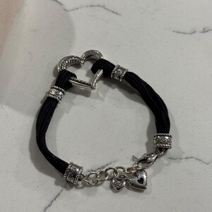 K&ouml;nnte beinhalten: Ein schwarzes Armband mit einem herzf&ouml;rmigen Silberverschluss und dekorativen silbernen Perlen. Das Armband hat eine Kette mit zwei kleinen Herzanh&auml;ngern. Das Design ist schlicht, aber elegant.