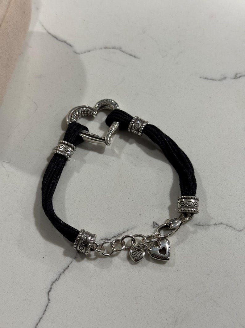 K&ouml;nnte beinhalten: Ein schwarzes Armband mit einem herzf&ouml;rmigen Silberverschluss und dekorativen silbernen Perlen. Das Armband hat eine Kette mit zwei kleinen Herzanh&auml;ngern. Das Design ist schlicht, aber elegant.