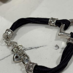 K&ouml;nnte beinhalten: Ein schwarzes Kordelarmband mit silberfarbenen Metallakzenten. Das Armband hat dekorative Verschl&uuml;sse und einen herzf&ouml;rmigen Anh&auml;nger. Das Armband liegt auf einer wei&szlig;en Oberfl&auml;che.