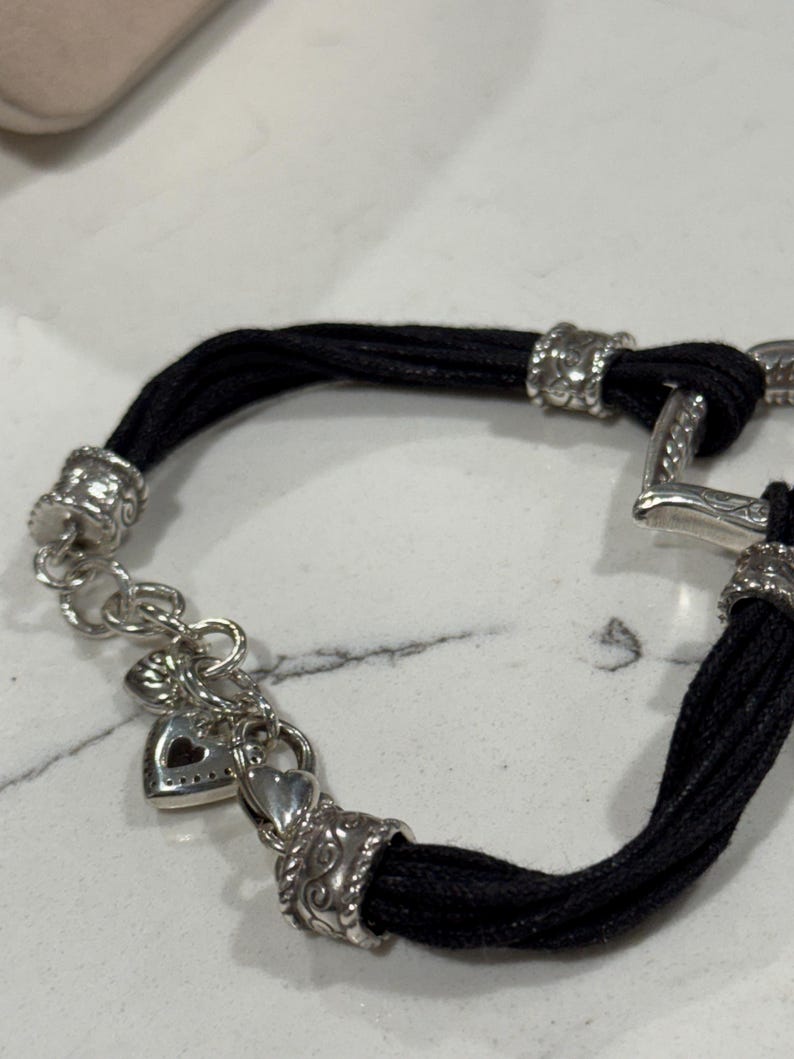 K&ouml;nnte beinhalten: Ein schwarzes Kordelarmband mit silberfarbenen Metallakzenten. Das Armband hat dekorative Verschl&uuml;sse und einen herzf&ouml;rmigen Anh&auml;nger. Das Armband liegt auf einer wei&szlig;en Oberfl&auml;che.