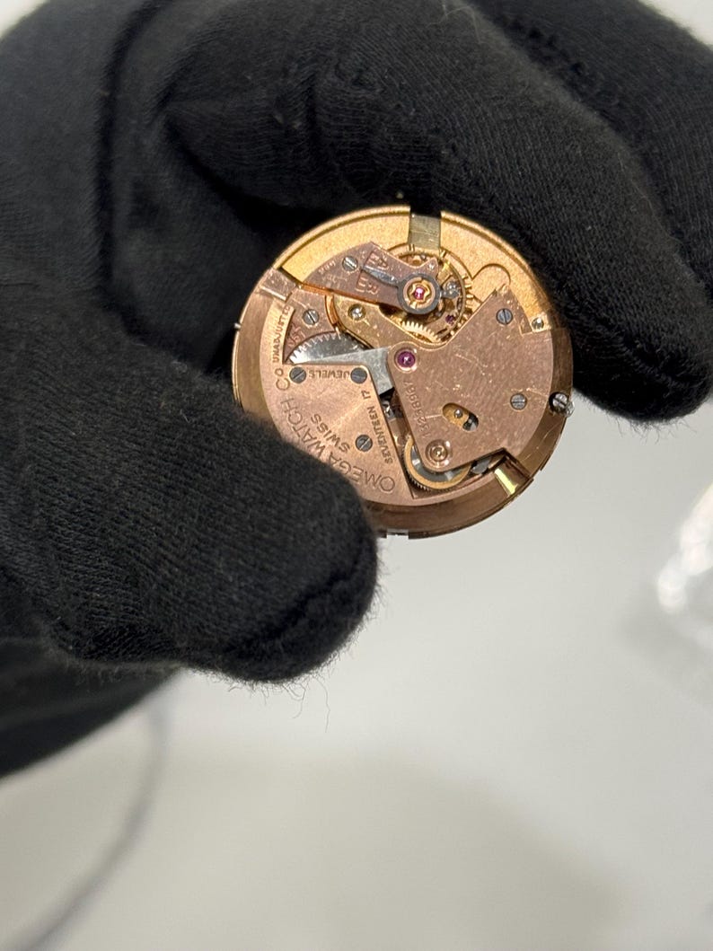 May include: Close-up of a watch movement with intricate gears and components. The watch mechanism is gold-toned with visible jewels and the text "OMEGA WATCH CO SWISS". The watch movement is held by a black gloved hand.