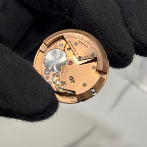 May include: A close-up of a gold-toned watch movement, revealing intricate gears and components. The text "OMEGA WATCH CO SWISS" and "SEVENTEEN 17 JEWELS" are visible. The watch mechanism is held by a black gloved hand.