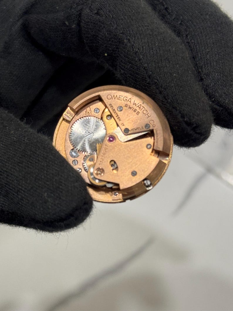May include: A close-up of a gold-toned watch movement, revealing intricate gears and components. The text "OMEGA WATCH CO SWISS" and "SEVENTEEN 17 JEWELS" are visible. The watch mechanism is held by a black gloved hand.