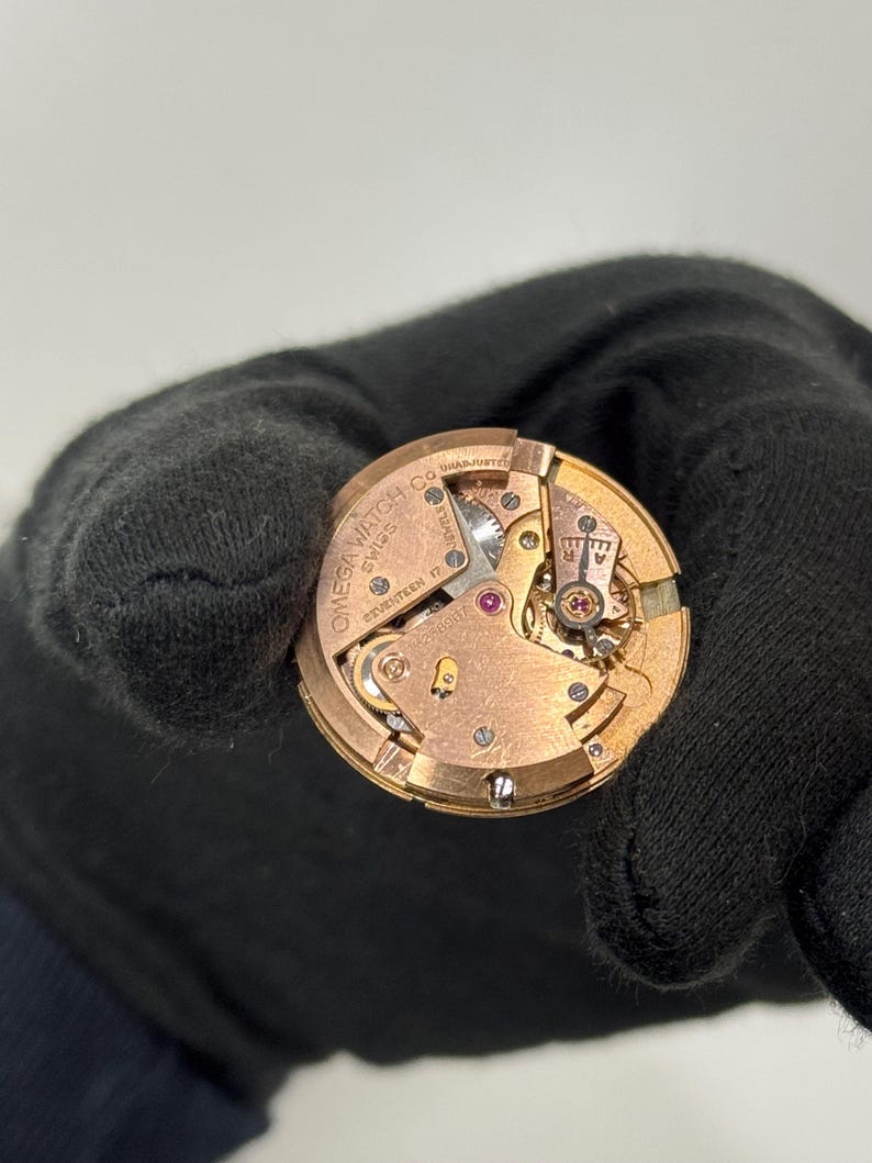 May include: A detailed view of a gold-coloured watch movement, showcasing the internal gears and mechanisms. The watch component is stamped with "OMEGA WATCH CO SWISS" and "SEVENTEEN 17 JEWELS". The image is set against a plain backdrop.