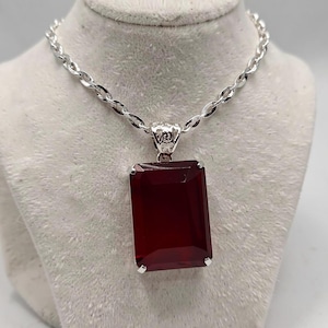 Könnte beinhalten: Eine silberne Halskette mit einem rechteckigen, tiefroten Anhänger. Der Anhänger ist in einen silbernen Rahmen gefasst und hängt an einer silbernen Kette. Die Halskette wird auf einer weißen Büste präsentiert.